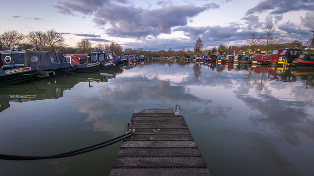 View from the end of a pontoon in a marina full of narrowboats under an unsettled cloudy sky.