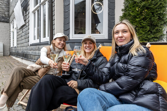 Three smiling women toast with sparkling wine outside cafe in Europe, enjoying friendship, travel and celebration. Candid lifestyle scene captures leisure, reunion, happiness and autumn atmosphere.