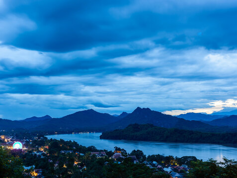 Dusk view of Luang Prabang, Laos, showing the Mekong River and dark mountains beneath a cloudy blue evening sky from Phousi Hill.