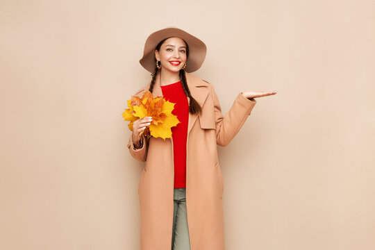 A beautiful young woman in an autumn fashionable outfit with shopping bags and round hat pointing aside at free space over beige background