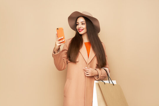 A stylish woman, dressed in fashionable autumn outfit including beige coat and hat, smiles while looking at her smartphone, holding several shopping bags against beige background