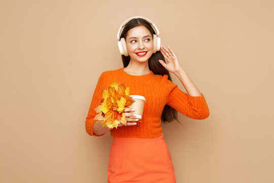 A beautiful young woman in a fashionable autumn outfit listens to music with wireless headphones. A girl in a stylish orange blouse poses on a beige background