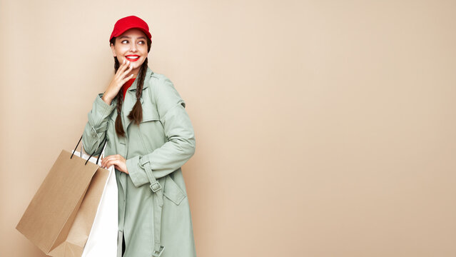 A beautiful young woman in an autumn raincoat shows shopping bags posing on a beige studio background