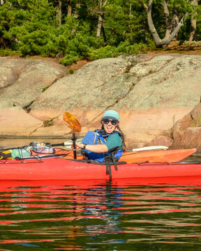 Playful young woman sticks out her tongue while sea kayaking in a red kayak beside granite shore with copy space on Georgian Bay, Ontario, Canada