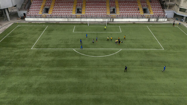 Aerial view of a football match in progress at a stadium in Lagos Island, featuring players on the green pitch, empty spectator seating, and clear markings on the field in Lagos, Lagos, Nigeria.