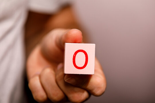 A hand extends forward, presenting a light pink wooden block with a red 'O' on it.