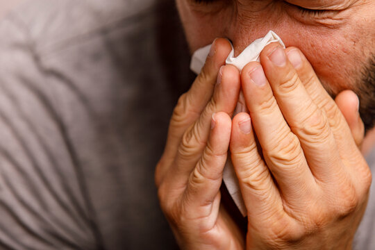 A close-up of a man blowing his nose with a tissue, indicating illness or allergies.