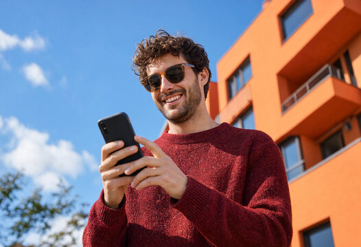 Cheerful man enjoys reading his phone outdoors near an orange building on a sunny day