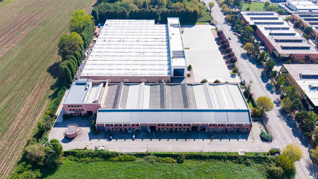 Aerial view of an industrial facility with large warehouses, flat roofs, and parking areas, situated near agricultural fields and roads in Milan, Lombardy, Italy.