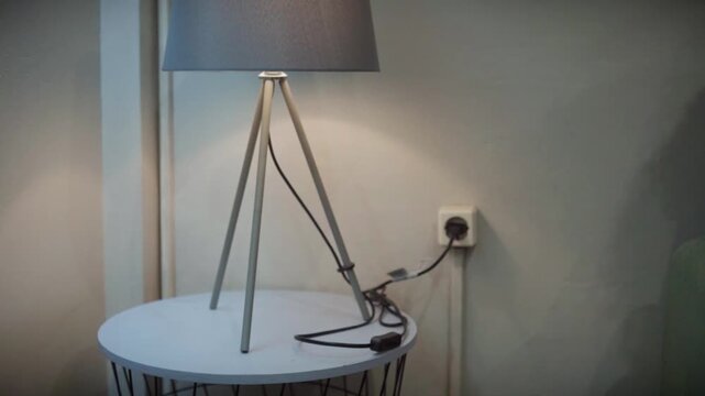 Hand plugging lamp into wall outlet. Soft light illuminates bedside table. Minimalist design adds calm atmosphere. Ideal for cozy home or bedroom scenes. Simple, quiet moment captured in dim room