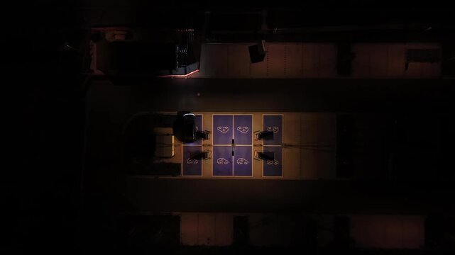 Single car parks among rows of illuminated blue electric vehicle charging spaces in USA, warm dim yellow lighting spreads across empty modern parking garage structure. Aerial drone top down rising