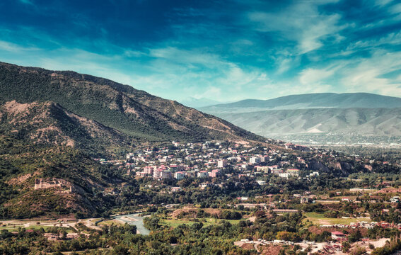 Scenic aerial view of historic Mtskheta town nestled in green valley surrounded by Caucasus mountains and winding river under blue cloudy sky in Georgia.
