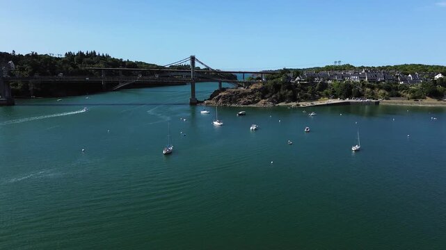 Wide cinematic drone shot over the Rance estuary, revealing Pont Saint-Hubert and a peaceful Breton village in the distance. Dozens of sailboats float across the shimmering blue-green water under a cl