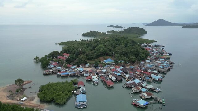 Aerial view of colorful stilt houses built over the water on Batam Island, surrounded by lush green hills and calm sea in Batam, Riau Islands, Indonesia.