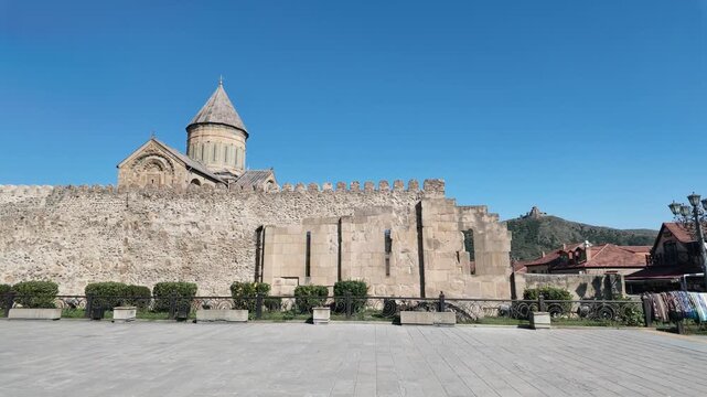 Video view of Svetitskhoveli Cathedral behind ancient fortified walls in Mtskheta, Georgia, with Jvari Monastery visible on a distant hill under clear blue sky. Svetitskhoveli Cathedral and Historic W