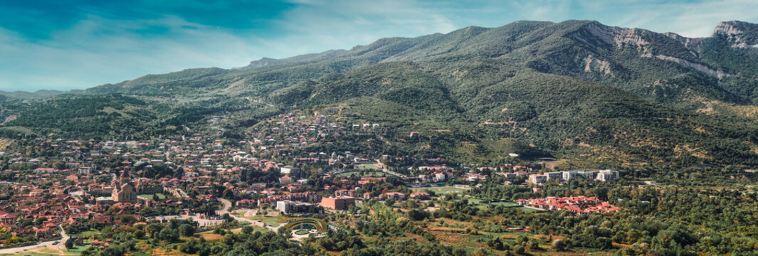 Wide aerial panorama of historic Mtskheta town surrounded by lush green Caucasus mountains under blue cloudy sky in Georgia. UNESCO heritage landmark view.