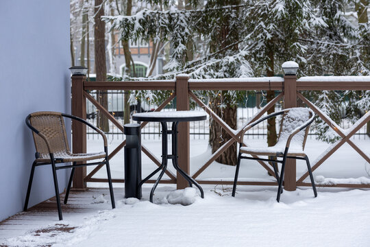 Snow covered outdoor cafe table and two chairs on wooden terrace with snowy trees in background in winter daylight