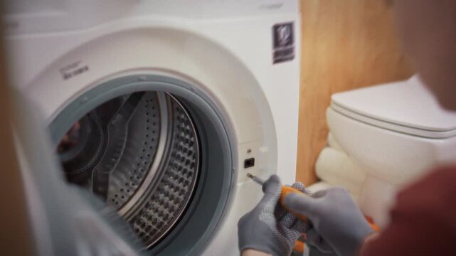 A technician in uniform and gloves works on a washing machine body with a toolbox and toilet visible in the bathroom background. Concept of calling a specialist for home appliance repair
