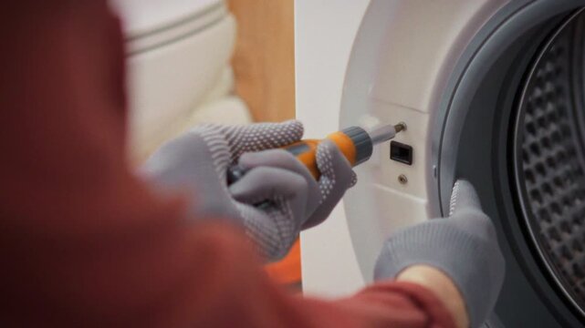 A technician in uniform carefully removes bolts from the body of a front-loading washing machine in a bathroom next to a toolbox. Concept of qualified home appliance repair services