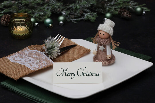 Festive Christmas table setting with white plate, rustic cutlery holder, cute wooden doll and a Merry Christmas card on a dark background.