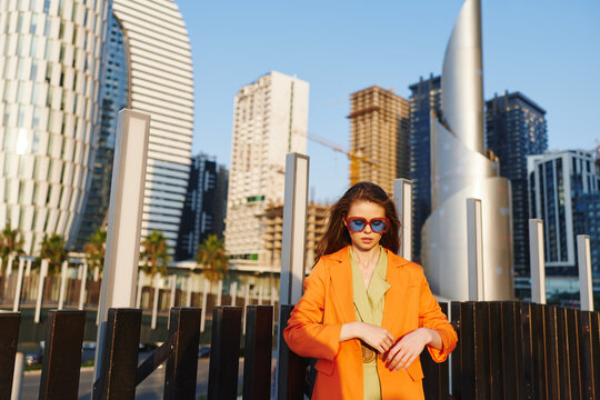 Stylish young woman in an orange blazer and blue mirrored sunglasses stands front-facing against futuristic skyscrapers. High fashion urban concept with modern architecture.