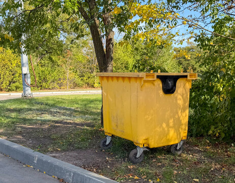 Large industrial yellow plastic waste container dustbin on wheels outdoors. Empty garbage bin skip for recycling sorting and municipal trash disposal management on a street near green trees. Urban. 