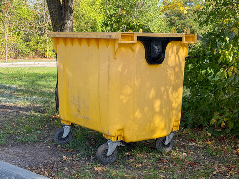 Large industrial yellow plastic waste container dustbin on wheels outdoors. Empty garbage bin skip for recycling sorting and municipal trash disposal management on a street near green trees. Urban. 