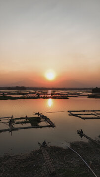 Aerial view of Cengklik Reservoir at sunset with Mount Merapi and Mount Merbabu in the distance, featuring bamboo fish farming structures and fishermen in Ngemplak, Central Java, Indonesia.