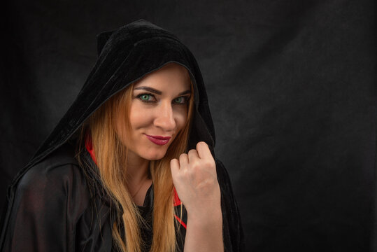 Young woman with captivating green eyes and long hair, wearing a mysterious black hooded cloak and smirking, posing with an intriguing expression against a dark background