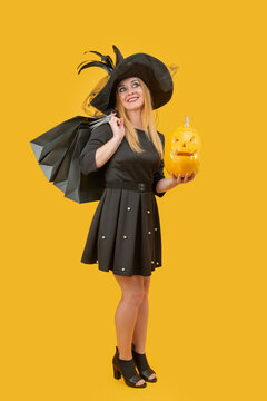 Woman in witch costume holding a carved jack o' lantern and black shopping bags, smiling during Halloween shopping against a bright yellow studio background, seasonal and joyful