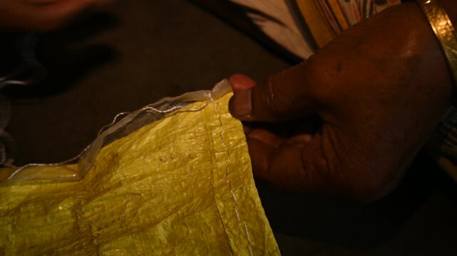 An elderly woman sews a rice sack by hand with thread and needle. Heavy-duty needles and thick plastic thread are commonly used tools for sealing bulk transport sacks. Wholesale markets (mandis)