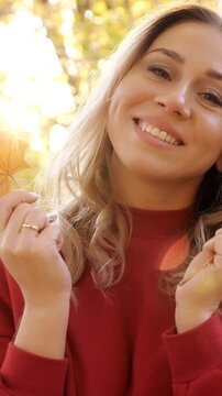 Smiling woman holds autumn leaves over face during portrait session. Woman in red sweater lowers colorful foliage to reveal her face, enjoying playful fall photography in warm golden light.