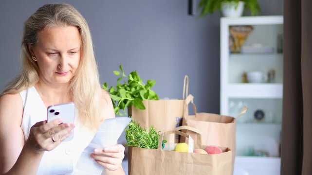 Mid adult woman comparing information on her smartphone and paper receipt beside groceries at paper bags at home in 4k real time footage. Grocery shopping, household expenses, and digital commerce.