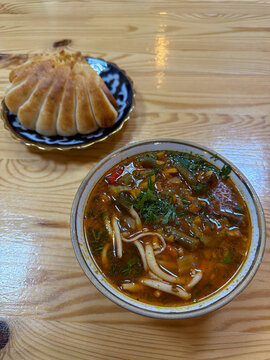 Traditional Central Asian laghman soup with handmade noodles, meat, and vegetables in a ceramic bowl served with a fried manti dumpling khoshan on a wooden table. Delicious oriental uzbek or uyghur. 