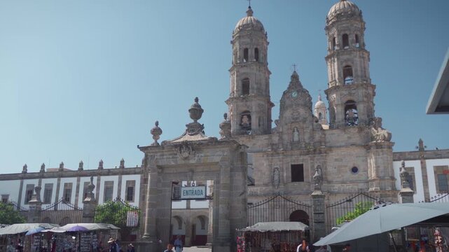 Basilica of Our Lady of Zapopan facade featuring colonial architecture, twin towers, central entrance, surrounding white walls with windows, and visitors walking under clear blue sky
