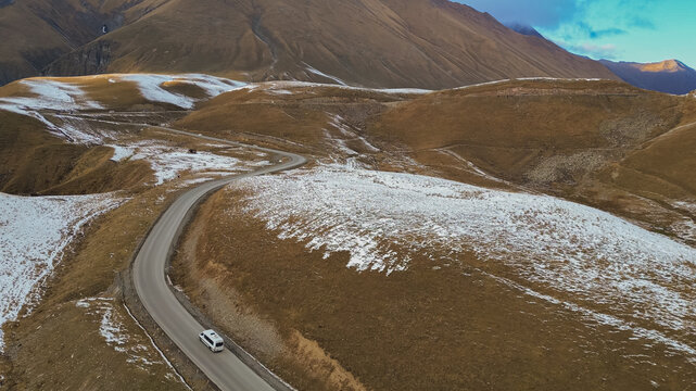 Horizontal drone view of white van driving along winding asphalt Georgian Military Road through snow dusted mountain pass in Georgia during late autumn
