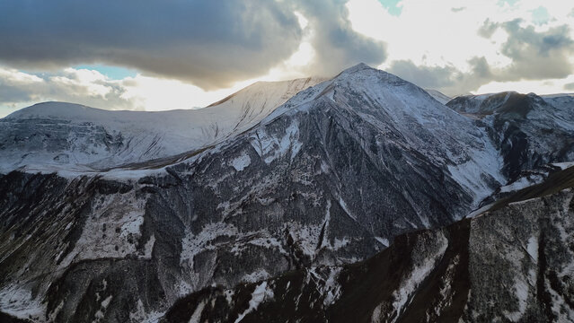 Horizontal drone view of majestic snow capped mountain peak touching dramatic clouds with sun rays breaking through in Georgia during late autumn