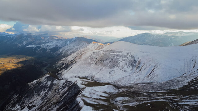 Horizontal aerial view of snow covered Greater Caucasus mountain range illuminated by sunlight under dramatic sky in Georgia during late autumn