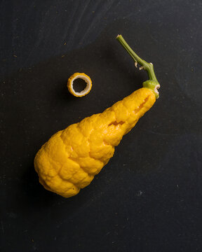 A single elongated yellow citrus fruit with a bumpy texture and a green stem, positioned next to a small circular slice on a dark slate surface