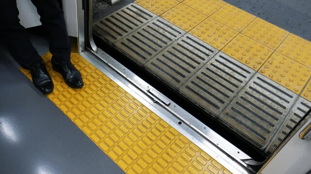 High angle shot of a commuter's feet standing inside a train by open sliding doors. Yellow tactile paving on the floor and automated platform screen gates visible at a modern subway station.