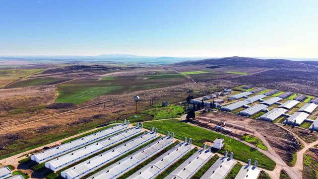 Sprawling poultry farm rows across rural valley. Wide aerial view captures barn rooftops, silos, water tower and distant hills.