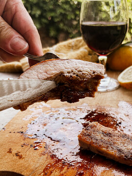 Person carving juicy grilled beef from an Argentine asado with a fork and knife, preparing a delicious gourmet meal with red wine and fresh lemons