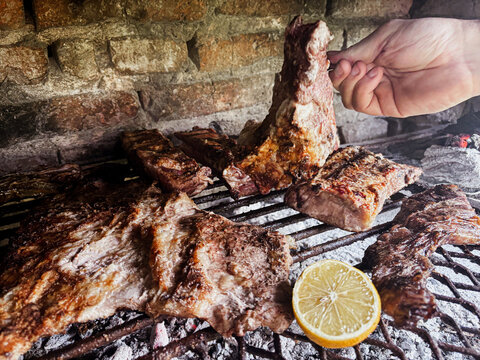 Hand turning sizzling beef ribs and various cuts of asado on a hot charcoal grill, creating a traditional Argentinian barbecue experience with a fresh lemon