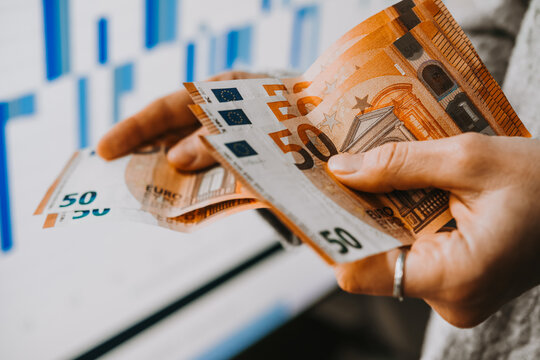 Hands holding euro currency in front of the stock exchange screen showing trading graphics.