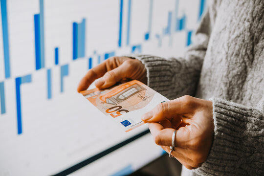 Hands holding euro currency in front of the stock exchange screen showing trading graphics.