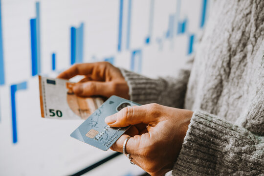 Hands holding euro currency and a bank card in front of the stock exchange screen showing trading graphics.