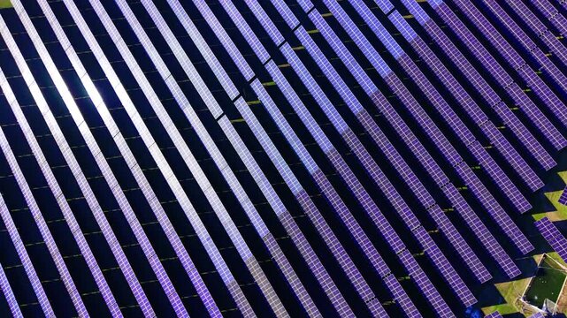 Vast solar panel array spans open field. Overhead drone of countless diagonal rows with bright reflective glare sections.