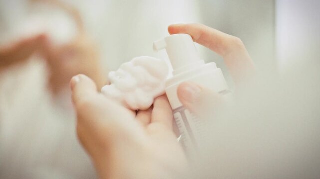 In a bright bathroom, a girl stands before a mirror, squeezing out a generous amount of white airy foam from a bottle for her facial cleansing routine, preparing her skin with a refreshing wash