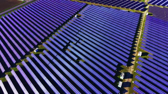 Morning light over solar panel installation. Aerial view reveals panel rows, small white sheds, access road and green field edges.