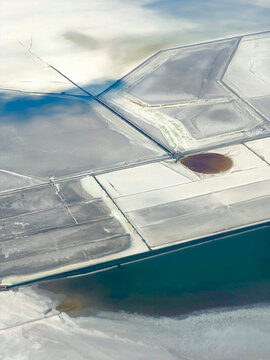 An aerial view of geometric salt evaporation ponds near the Great Salt Lake, Utah, in pale white and gray tones bordering a channel of dark blue-green open water.